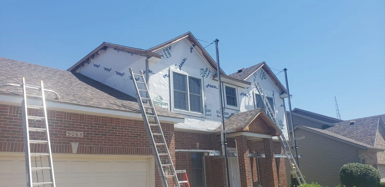 Brick home with ladders against a house-wrapped gable during siding work